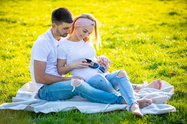 Lyckligt par väntar baby, gravid ung kvinna med make sitter på grönt gräs och håller baby stövlar, unga familj och New Life Concept — Stockfoto