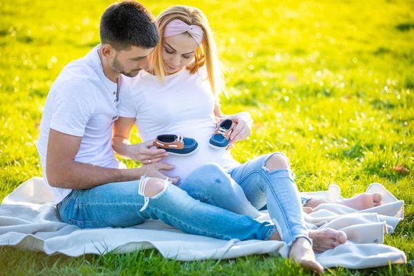 Pareja feliz esperando bebé, mujer joven embarazada con marido sentado en la hierba verde y la celebración de botas de bebé, familia joven y nuevo concepto de vida —  Fotos de Stock