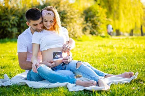 Pareja feliz esperando bebé, mujer joven embarazada con marido sentado en la hierba y la celebración de imágenes de ultrasonido, familia joven y nuevo concepto de vida —  Fotos de Stock