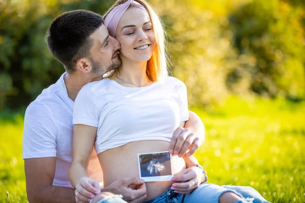 Feliz casal esperando bebê, grávida jovem com marido sentado na grama e segurando ultra-som imagens, jovem família e novo conceito de vida — Fotografia de Stock