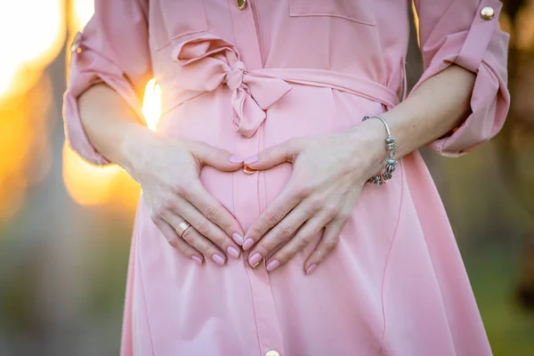 Händerna på gravida kvinnan på hennes mage i parken vid soluppgången med en varm bakljus i bakgrunden, New Life Concept — Stockfoto