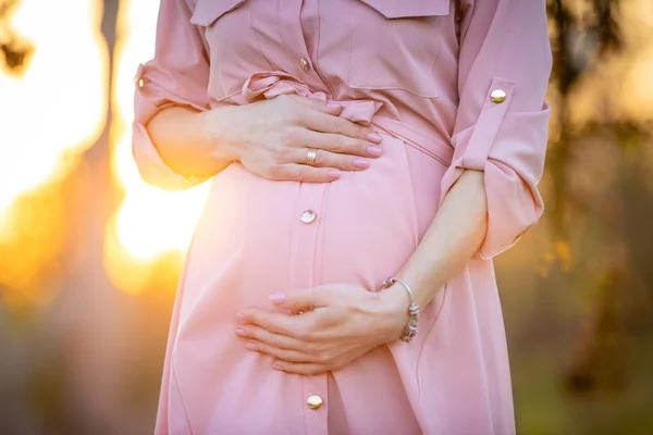 Manos de mujer embarazada sobre su vientre en el parque al amanecer con una cálida luz de fondo, nuevo concepto de vida —  Fotos de Stock