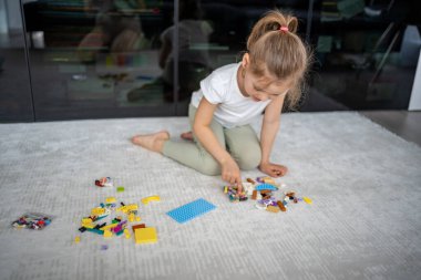 Küçük kız yerde oturuyor ve oturma odasındaki yapı taşlarıyla oynuyor. Erken gelişim kavramı, yaratıcılık ve ev ortamında odaklanmış oyun. Yüksek kalite fotoğraf