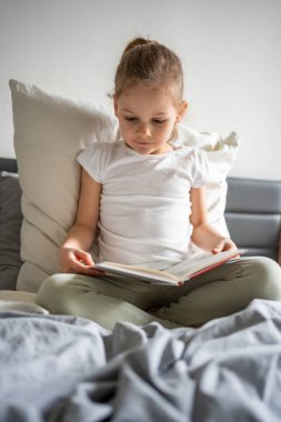 Küçük kız ailesinin yatağında oturuyor ve yatak odasında kitap okuyor. Sessiz zaman kavramı, konsantrasyon ve erken yaşta okuma aşkı. Yüksek kalite fotoğraf