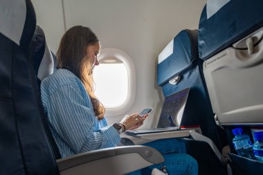 Uçuş sırasında dizüstü bilgisayarda çalışan genç bir kadın, uçak penceresinin yanında oturuyor. Uzaktan çalışma, seyahat verimliliği ve dijital yaşam tarzı devam ediyor. 