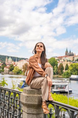 Sırtında küçük bir sırt çantası olan genç bir kadın Prag Kalesi 'nin arka planında bulanık bir şekilde taş korkulukta oturuyor. Tek başına seyahat, barışçıl keşif ve tarihi Avrupa şehirlerini keşfetme kavramı. 