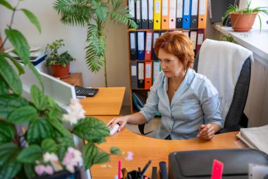 Arkasında dosyalar, belgeler ve bitkiler olan dağınık bir ofis masasında çalışan bir kadın. Günlük iş akışı, evrak işleri ve gerçek idari çalışma ortamı kavramı. 