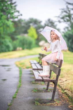 Little girl sitting on a park bench with an umbrella in her hands during light rain. Calm and reflective pause surrounded by nature and soft weather. High quality photo
