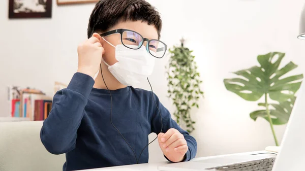 Online learning & Social distancing, Cute Asian little boy put on headphone to study online lessons from computer laptop at home due to Covid-19 pandemic, lockdown and school break / closure.