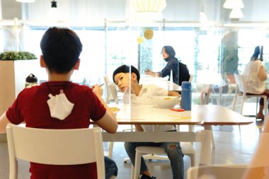 Cute little boy talk to his brother who sit on other side of table in food court with clear divider / barrier on table. New normal, Social & physical distancing during Covid-19 pandemic  clipart