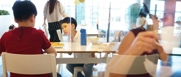 Cute little boy talk to brother who sit on the other side of table with clear divider / barrier on table in restaurant. New normal, Social or physical distancing during Covid-19 pandemic concept.