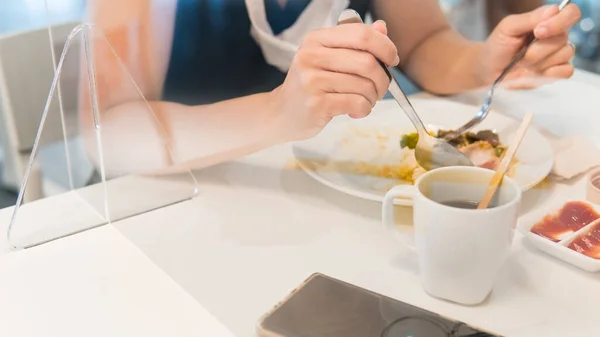 Kadının elleri, boynunda tıbbi maskeyle tek başına yemek yerken akrilik ayırıcının / bariyerin diğer tarafında otururken görülüyor. Covid-19 salgını sırasında yeni normal & sosyal uzaklık