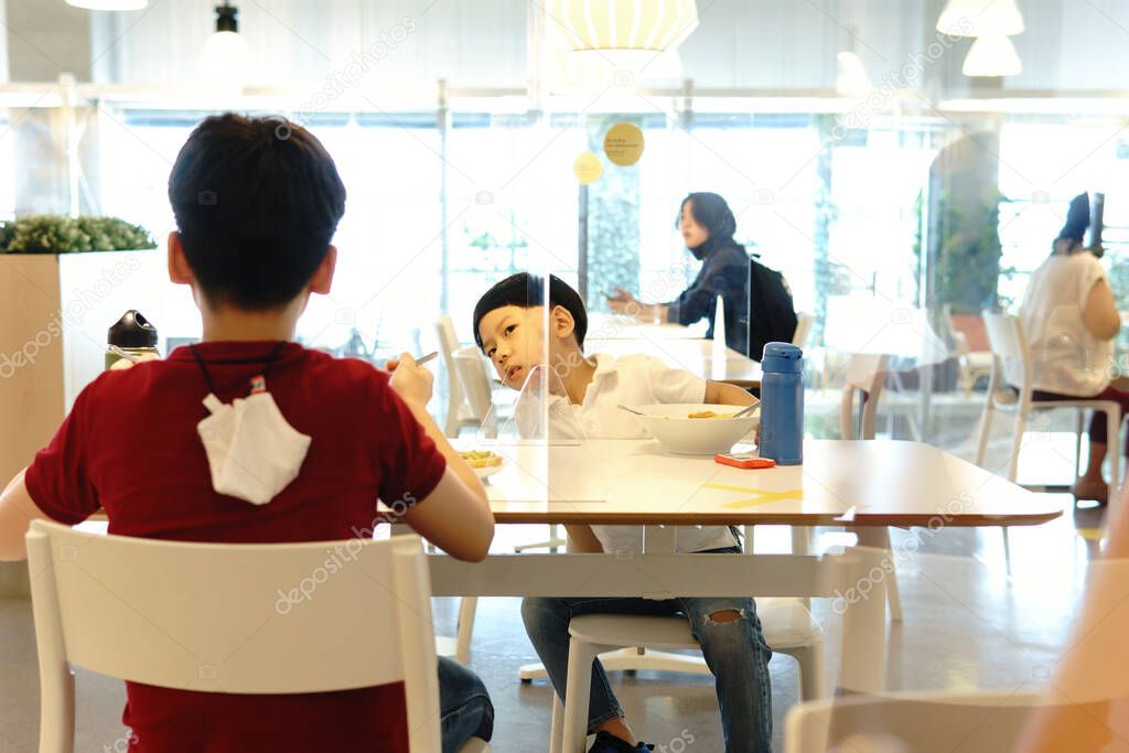 Cute little boy talk to his brother who sit on other side of table in food court with clear divider / barrier on table. New normal, Social & physical distancing during Covid-19 pandemic 