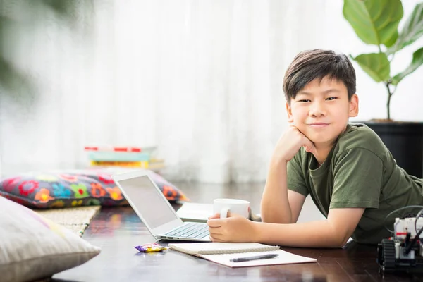 Smart looking Asian preteen boy lying on floor, smile with confidence and happiness, studying online learning at home due to Covid-19 pandemic, city lockdown and social distancing measures