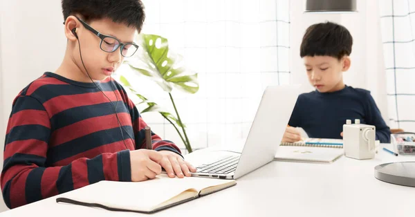 İki zeki Asyalı çocuk ders çalışıyor ve online dersler dinliyor. Covid-19 salgınının karantinası sırasında evde deftere yazıyorlar. Yeni normal, sosyal uzaklık, çevrimiçi öğrenme ve ev okulu kavramı