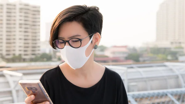 Sosyal mesafeli, güzel Asyalı kadın tıbbi yüz maskesi takıyor, sosyal medya uygulamasını kontrol ediyor ve Covid-19 salgını sırasında akıllı telefon üzerinde çalışıyor..