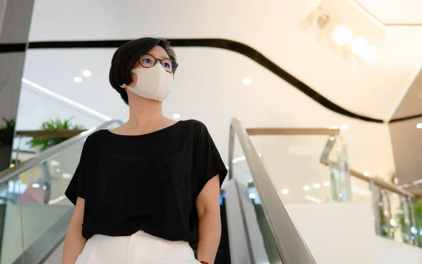 Güzel orta yaşlı Asyalı kadın yürüyen merdivende duruyor, virüs, salgın, grip salgını ve boş alışveriş merkezlerinde salgın hastalıklardan korunmak için tıbbi maske takıyor..