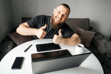 Kameraya bakan genç mutlu grafik tasarımcısı baş parmaklarıyla hareket gösteriyor. Evdeki işyerinde. Uzak çalışma.