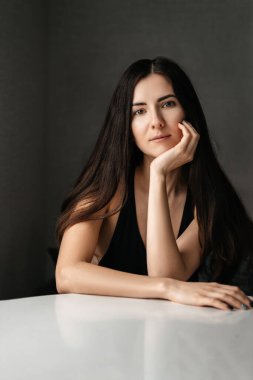 Genç, güzel, siyah saçlı bir kadın masaya oturmuş, elini uzatıp kameraya bakıyor.