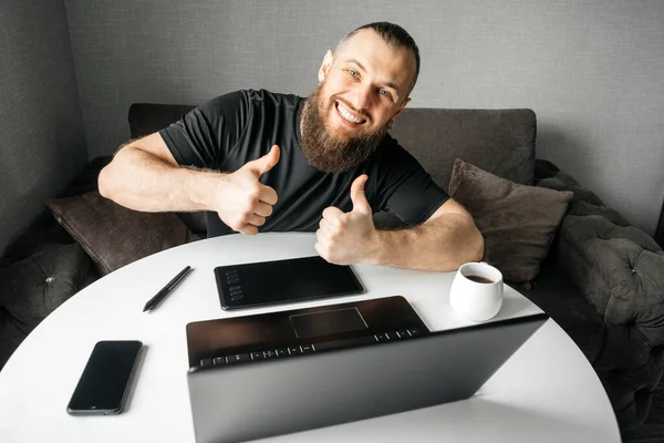 Kameraya bakan genç mutlu grafik tasarımcısı baş parmaklarıyla hareket gösteriyor. Evdeki işyerinde. Uzak çalışma.