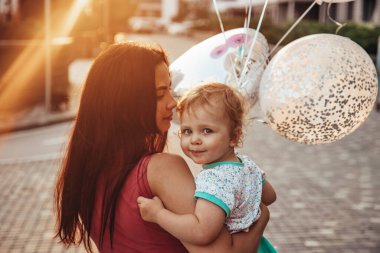 Annesinin kollarında mavi gözlü küçük bir çocuk. Balonlar ve güzel günbatımı.