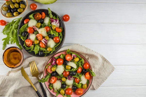 Beyaz ahşap zemin üzerinde kavun, domates ve zeytinli iki porsiyon salata. Metin için boş alan.