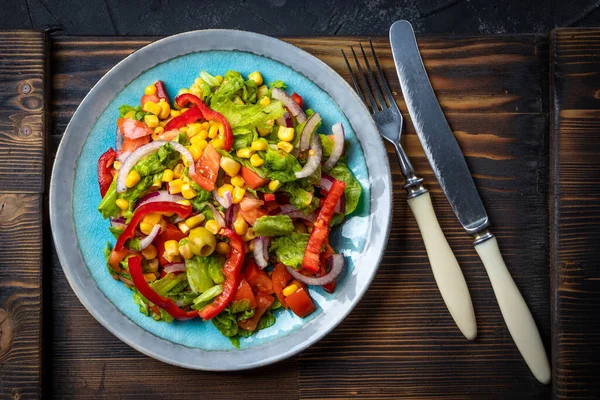 Fresh salad with corn, tomato and pepper in a plate on a wooden background, top view.