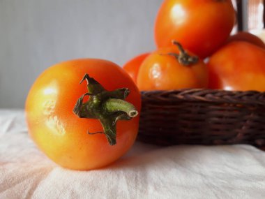 Olgun domatesler portakaldır. Beyaz kumaş tabanlı. Arka planda küçük bir hasır sepette domatesler var.