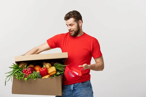 Delivery Concept: Handsome delivery man is holding a heavy grocery box isolated over grey background.
