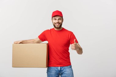 Neşeli, genç, yakışıklı teslimatçı elinde bir karton kutu ve gri arka planda dikilirken parmağını kaldırıyor.