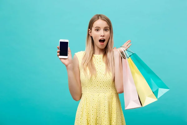 Portrét mladá atraktivní žena s nákupní tašky ukazuje obrazovka telefony přímo do fotoaparátu. Izolované na modrém pozadí — Stock fotografie