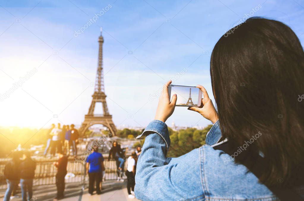 Woman tourist taking photo by phone near the Eiffel tower in Paris under sunlight and blue sky. Famous popular touristic place in the world.