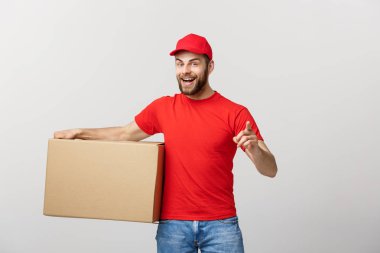 Neşeli, genç, yakışıklı teslimatçı elinde bir karton kutu ve gri arka planda dikilirken parmağını kaldırıyor.
