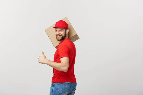 Joven y alegre repartidor guapo sosteniendo una caja de cartón y mostrando su pulgar hacia arriba mientras está de pie sobre un fondo gris —  Fotos de Stock