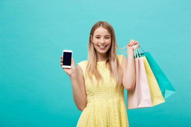 Alışveriş torbaları ile genç çekici kadın portre telefon ekranına doğrudan kamera gösterir. Üzerinde mavi arka plan izole