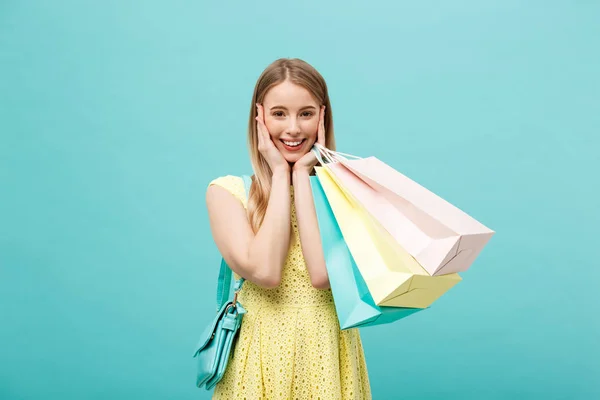 Portréja egy izgatott gyönyörű lány visel a sárga ruha, bevásárló táskák, elszigetelt, felett kék háttér gazdaság. — Stock Fotó