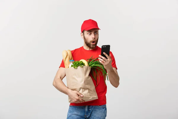 Доставщик держит бумажный пакет с едой и шоком или злится на что-то в мобильном телефоне на белом фоне — стоковое фото