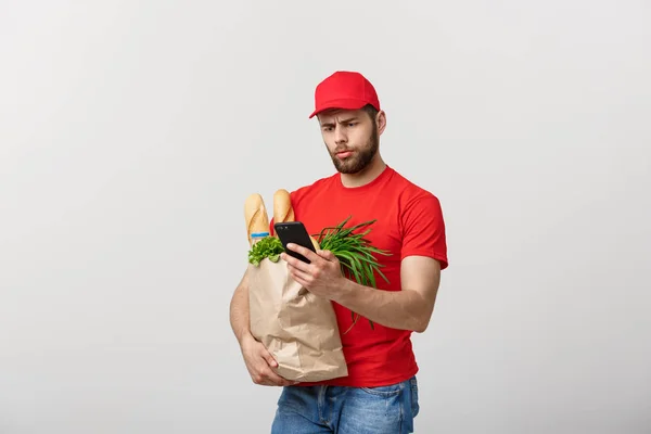 Entrega hombre sosteniendo bolsa de papel con comida y shock o enojado con algo en el teléfono móvil sobre fondo blanco —  Fotos de Stock