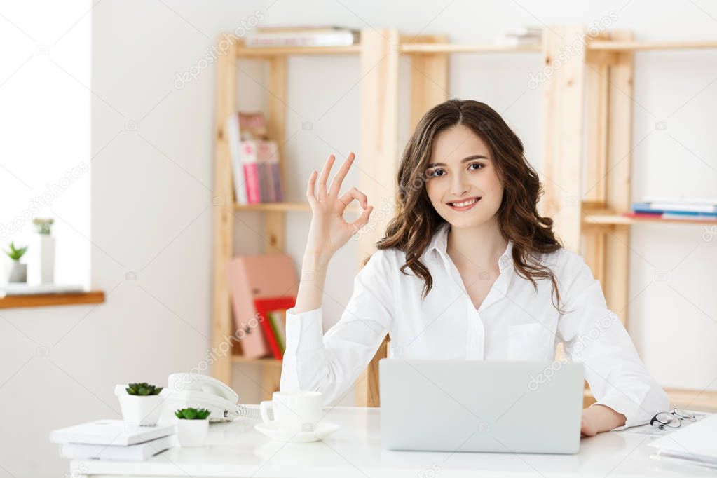 Young business woman showing ok sign sitting in the modern office with labtop.
