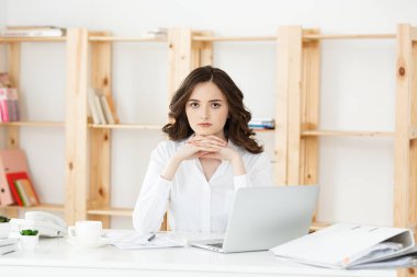 Modern bir ofis masasında dizüstü bilgisayarla çalışan ve bir şeyler düşünen çekici genç bir kadın..