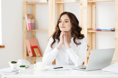Modern bir ofis masasında dizüstü bilgisayarla çalışan ve bir şeyler düşünen çekici genç bir kadın..