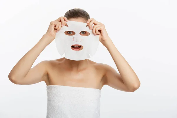 Mladá dívka s obličejovou maskou dívá na kameru na bílém pozadí. Kosmetická procedura. Kosmetické lázně a kosmetologie. — Stock fotografie