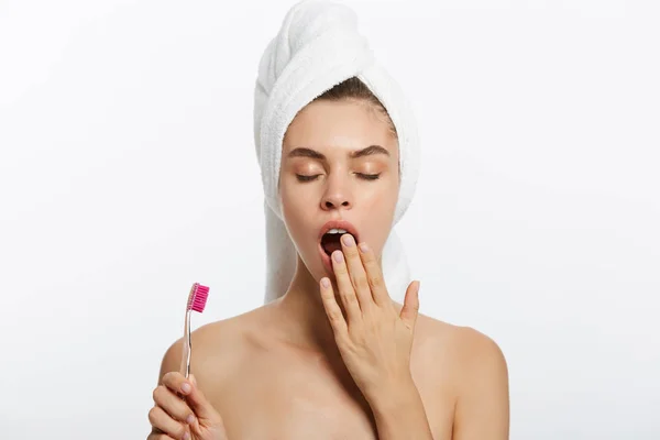 Senny młoda kobieta z biały ręcznik jest ziewanie i trzymając szczoteczkę do zębów na białym tle. — Zdjęcie stockowe