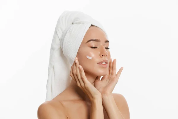 Primer plano retrato de belleza de una sonriente mujer semidesnuda con toalla en el pelo aplicando crema facial aislada sobre fondo blanco . —  Fotos de Stock