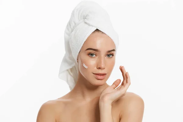 Primer plano retrato de belleza de una sonriente mujer semidesnuda con toalla en el pelo aplicando crema facial aislada sobre fondo blanco . —  Fotos de Stock