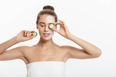 Yüzünde balçık maskesi olan genç bir kadın beyaz arka planda salatalık dilimleri tutuyor.