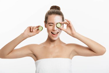 Yüzünde balçık maskesi olan genç bir kadın beyaz arka planda salatalık dilimleri tutuyor.