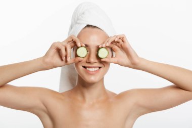 Bir gülümseyen güzel yarı çıplak kadın güzellik portresi kadar salatalık dilimleri tutarak ve beyaz arka plan üzerinde izole kamera bakarak saçlarını sarılı bir havlu ile kapatın