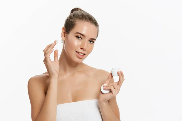 Wanita muda cantik menerapkan perawatan krim kosmetik di wajahnya terisolasi di latar belakang putih — Stok Foto