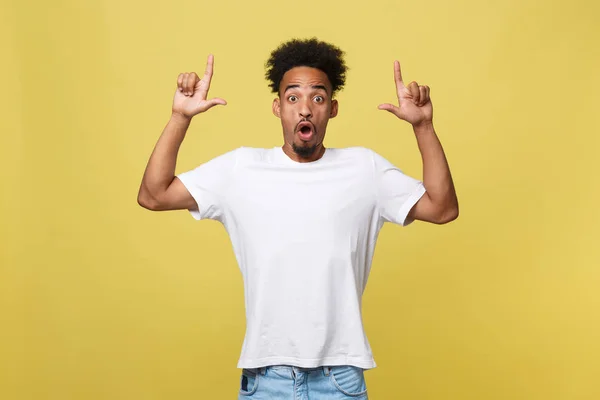 Förvånad ung African American man klädd i casual vit skjorta att ha glada fascinerad blick, pekar pekfingret på kopia utrymme på gyllene gul bakgrund för din text eller reklaminnehåll — Stockfoto